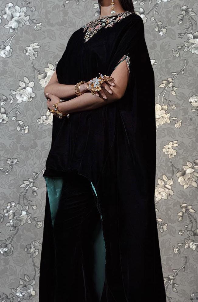 A person wearing a long black cape with gold embellishments and jewelry poses against a gray floral background.