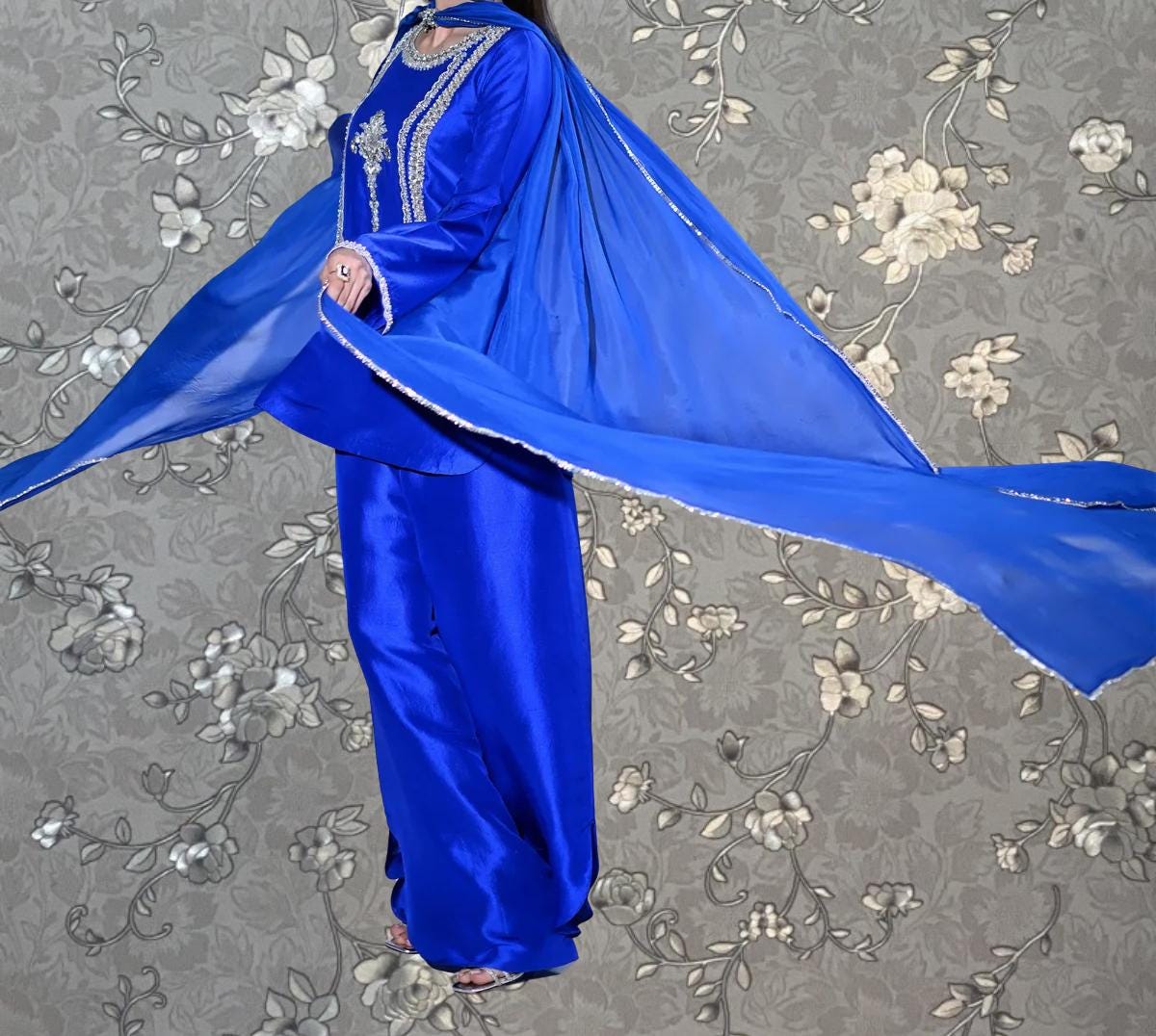 A person wearing a blue dress with a long flowing cape, standing against a gray floral background.