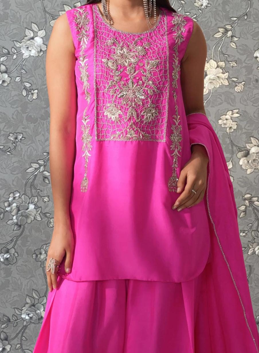 A woman wearing a bright pink dress with intricate embroidery stands against a gray floral background.