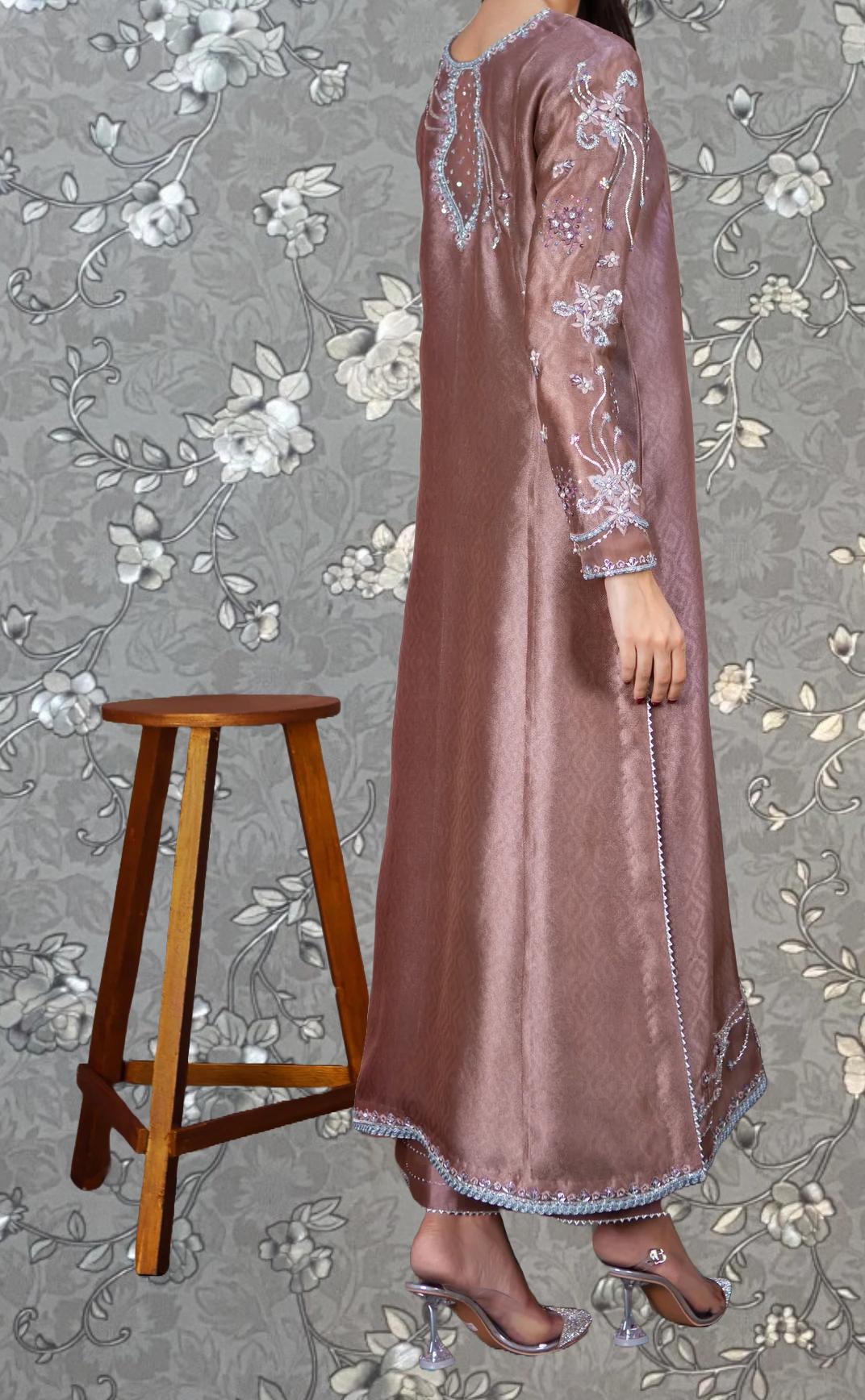 A woman wearing a long, pink dress with intricate embroidery stands next to a wooden stool against a gray floral background.