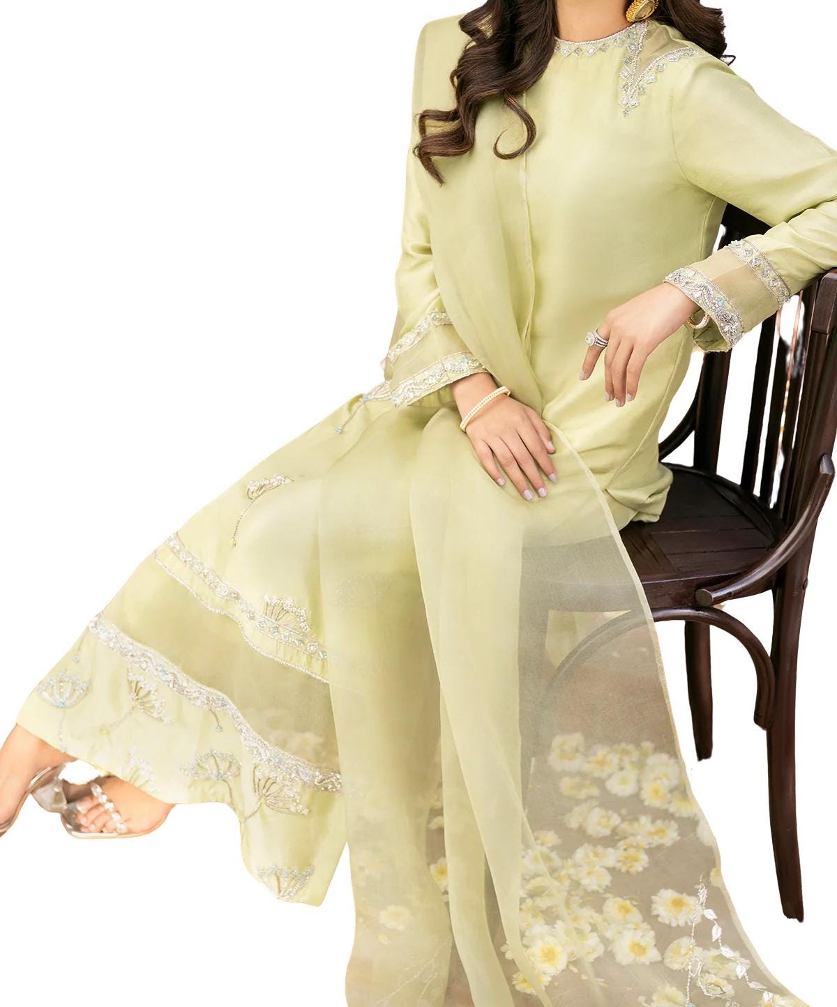 A woman wearing a long, flowing yellow dress with intricate embroidery sits on a wooden chair.