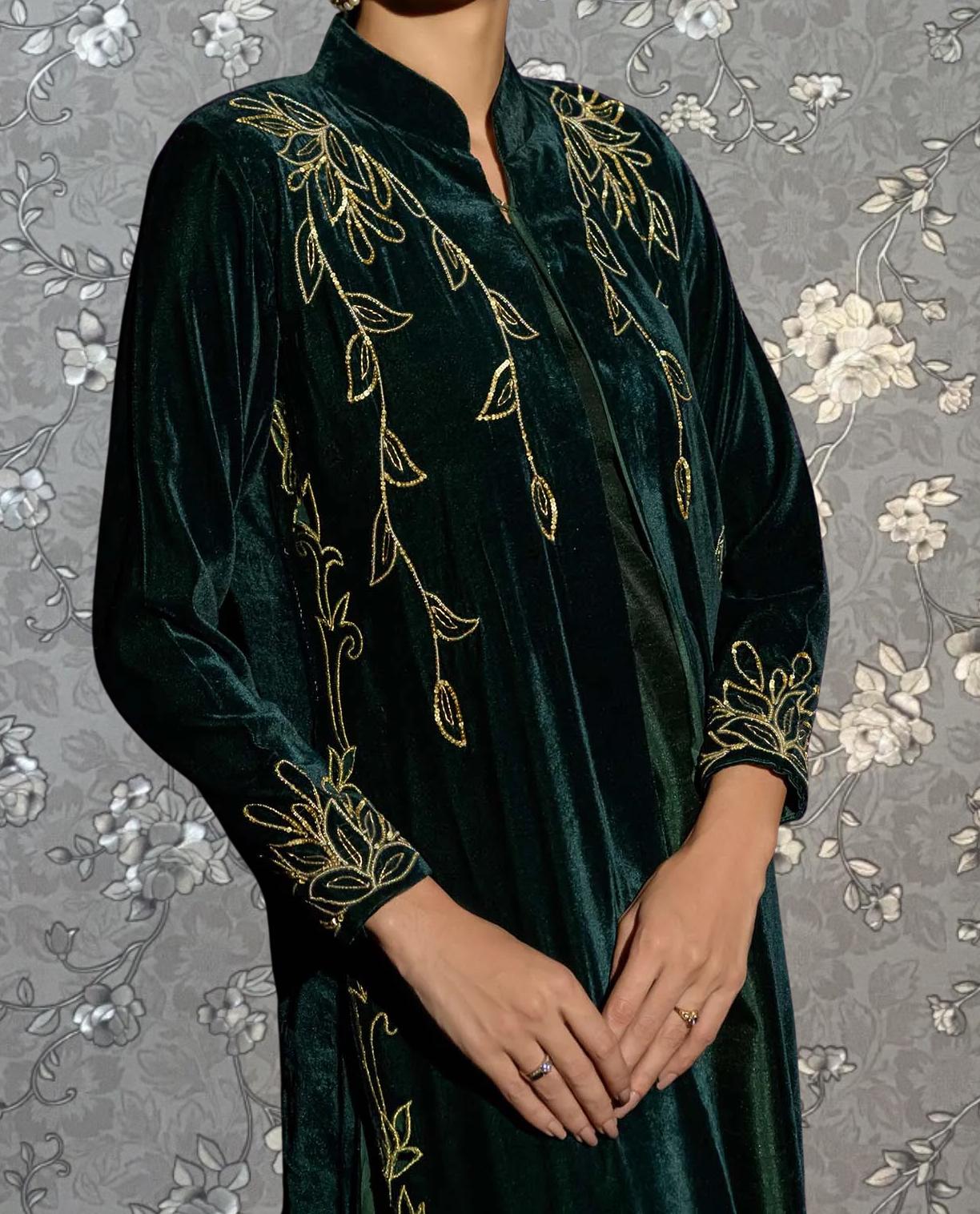 A person wearing a long, green velvet dress with gold embroidered leaf designs on the sleeves and collar.