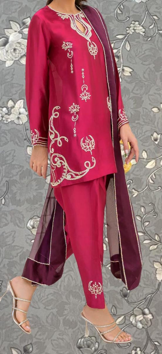 A person wearing a vibrant red and purple outfit with intricate embroidery, standing against a gray floral background.
