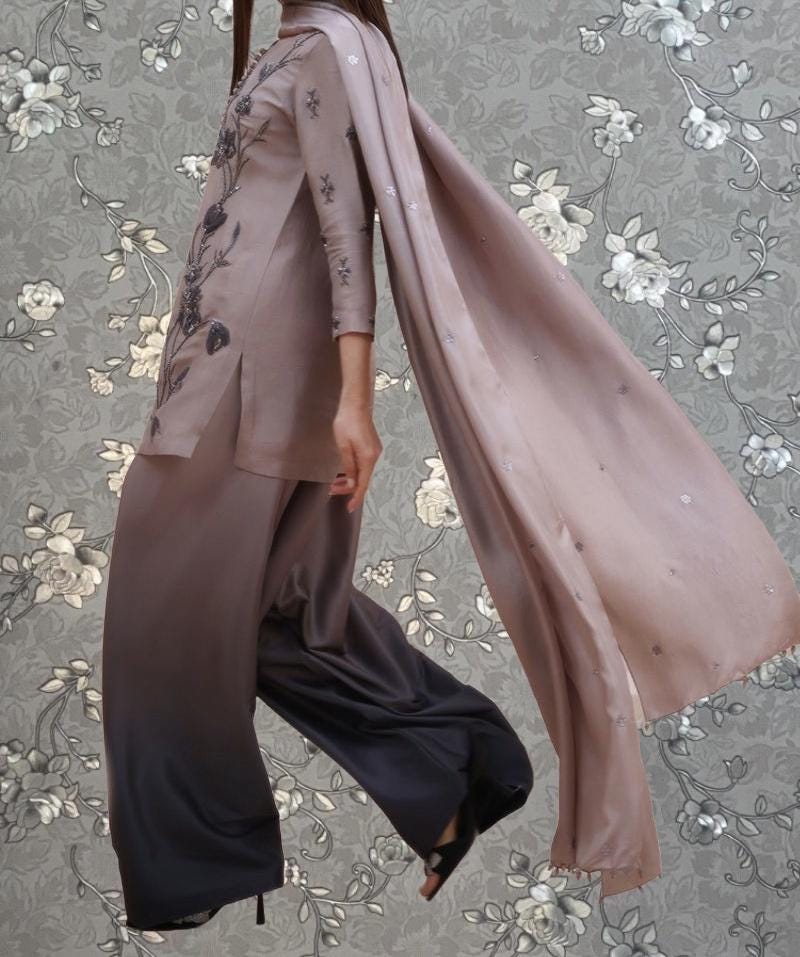 A person wearing a long, flowing pink dress with floral embroidery, standing against a gray floral background.