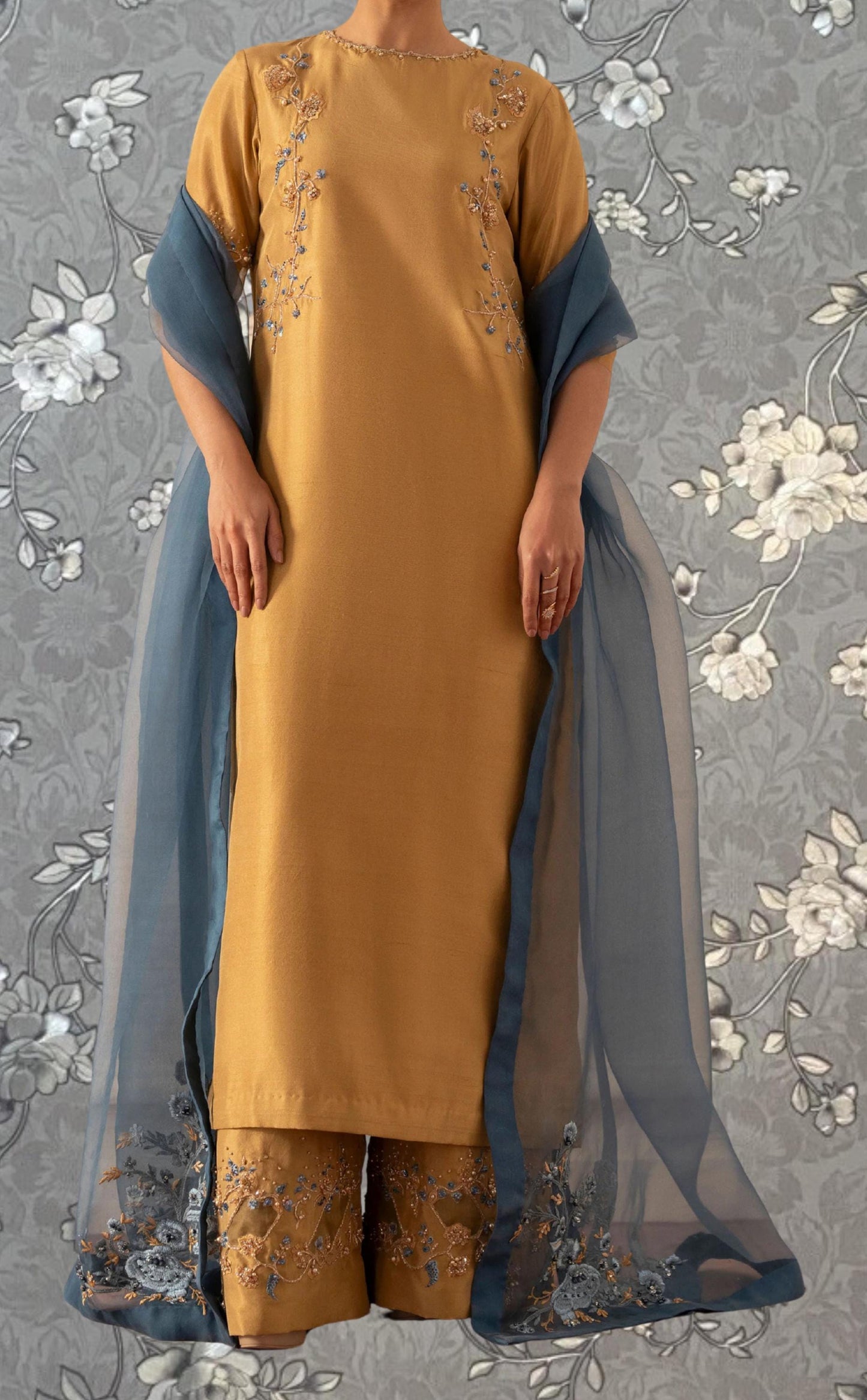 A person wearing a yellow dress with blue embroidered details and a matching blue shawl stands against a gray floral background.