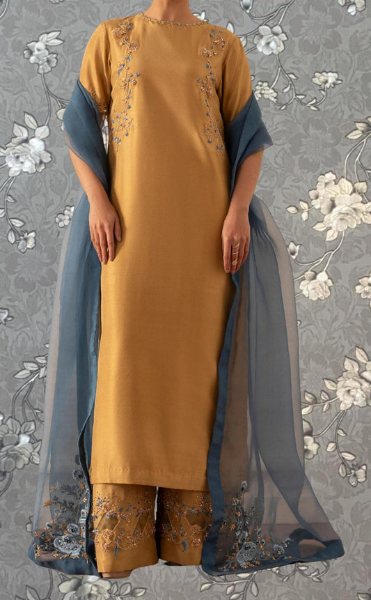 A person wearing a yellow dress with blue embroidered details and a matching blue shawl stands against a gray floral background.