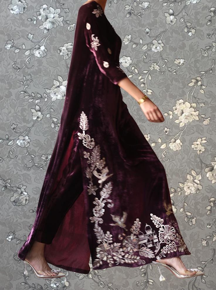 A person wearing a long, burgundy velvet dress with intricate floral embroidery on the sleeves and hem.