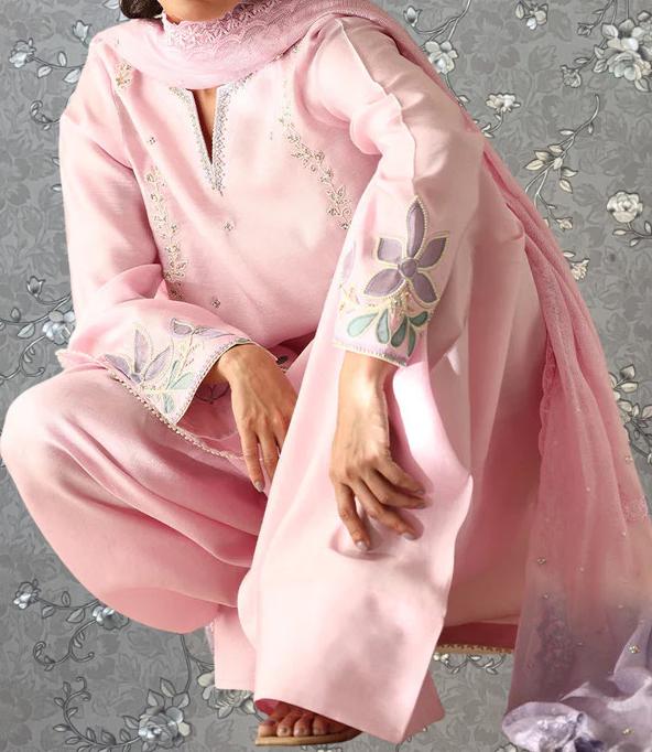 A woman wearing a pink dress with floral embroidery poses against a gray floral background.