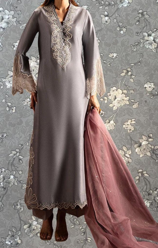 A woman wearing a gray dress with intricate embroidery stands against a gray floral background.