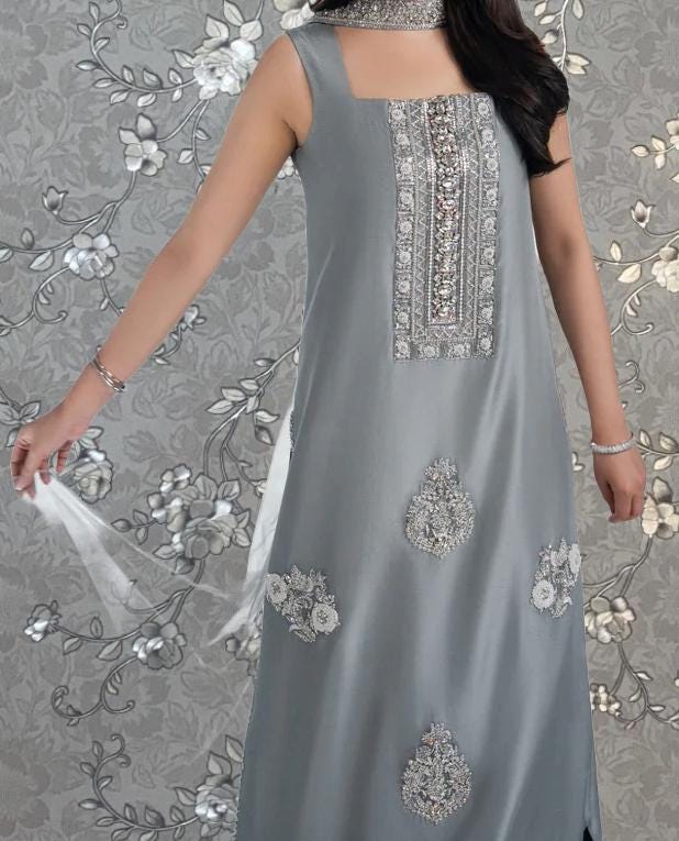 A woman wearing a gray dress with intricate embroidery and floral designs, posing against a gray floral background.
