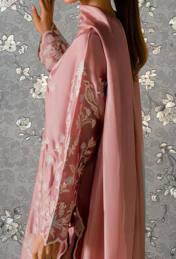 A woman wearing a pink dress with floral embroidery stands in front of a gray floral wallpaper.