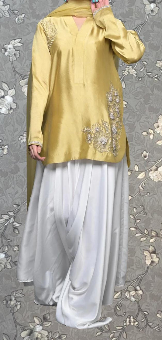 A person wearing a gold and white dress with floral embroidery, standing against a gray floral background.