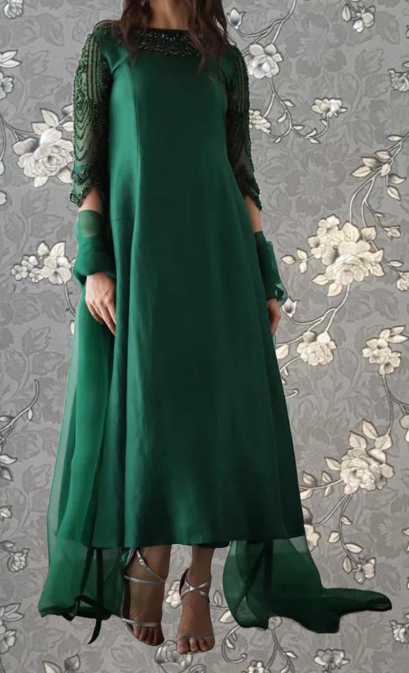 A woman wearing a long, green dress with lace sleeves and a flowing skirt stands against a gray floral background.