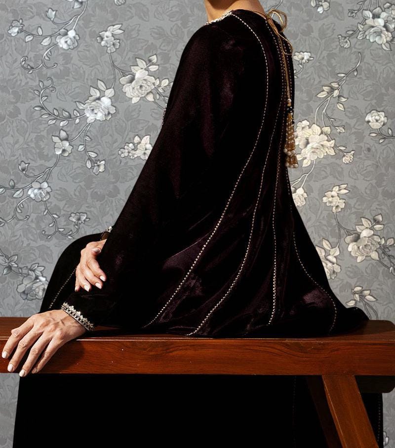 A person wearing a long black cape is sitting on a wooden bench against a gray floral wallpaper background.