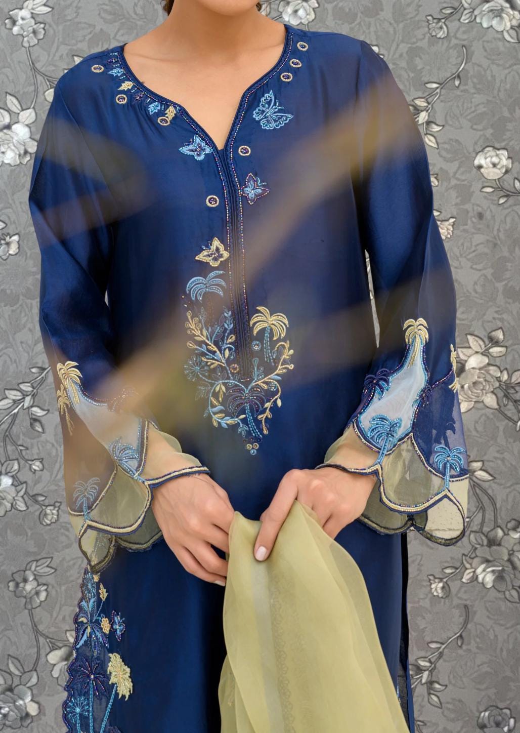 A woman wearing a blue dress with intricate embroidery and floral patterns, holding a yellow fabric in her hands.
