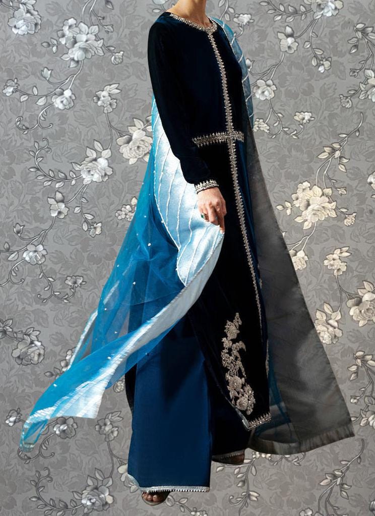 A person wearing a long black dress with a blue and white patterned shawl, standing against a gray floral background.