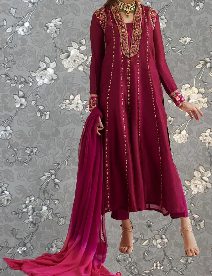 A woman wearing a long, burgundy dress with intricate embroidery and gold accents, standing against a gray floral background.
