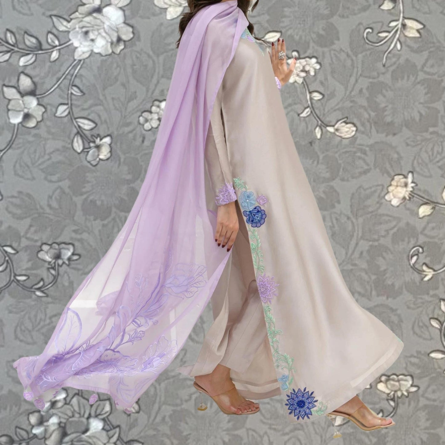 A woman wearing a long, flowing dress with a purple and blue floral pattern on the side, standing against a gray floral background.