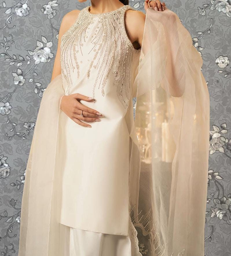 A woman wearing a white dress with a beaded bodice and a sheer, flowing skirt stands against a gray floral background.