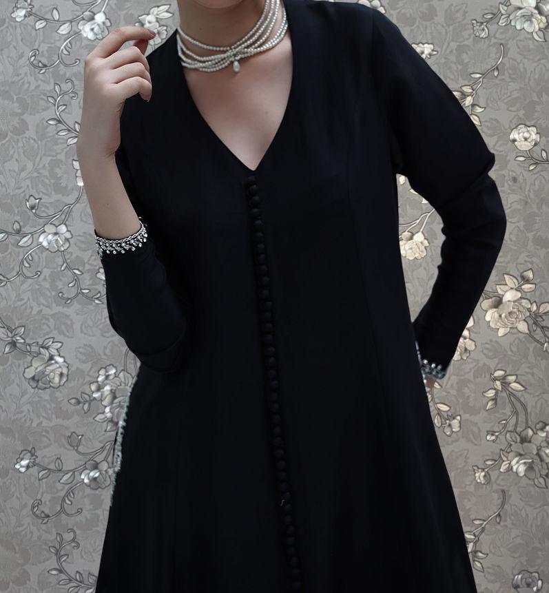 A woman wearing a black dress with a v-neck and long sleeves, accessorized with a pearl necklace and bracelets.
