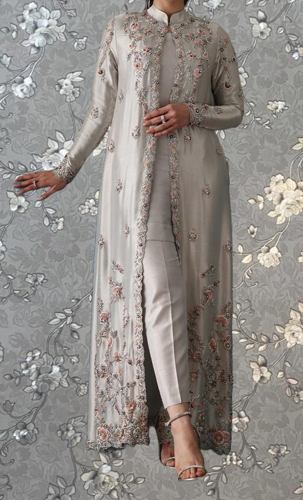 A woman wearing a long, gray dress with intricate floral embroidery stands against a gray floral background.