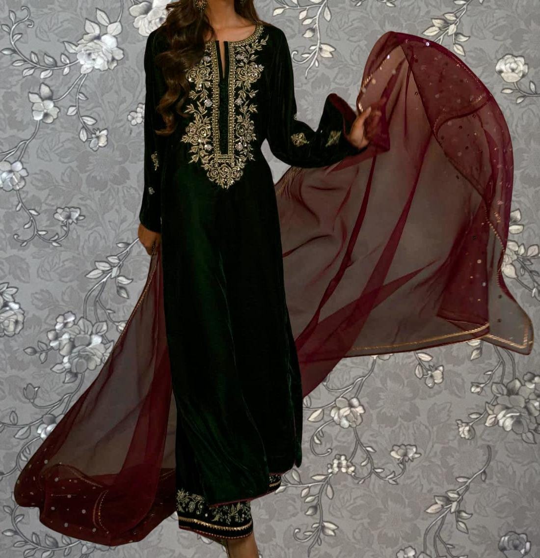 A woman wearing a long, green dress with gold embroidery and a red shawl is standing against a gray floral background.