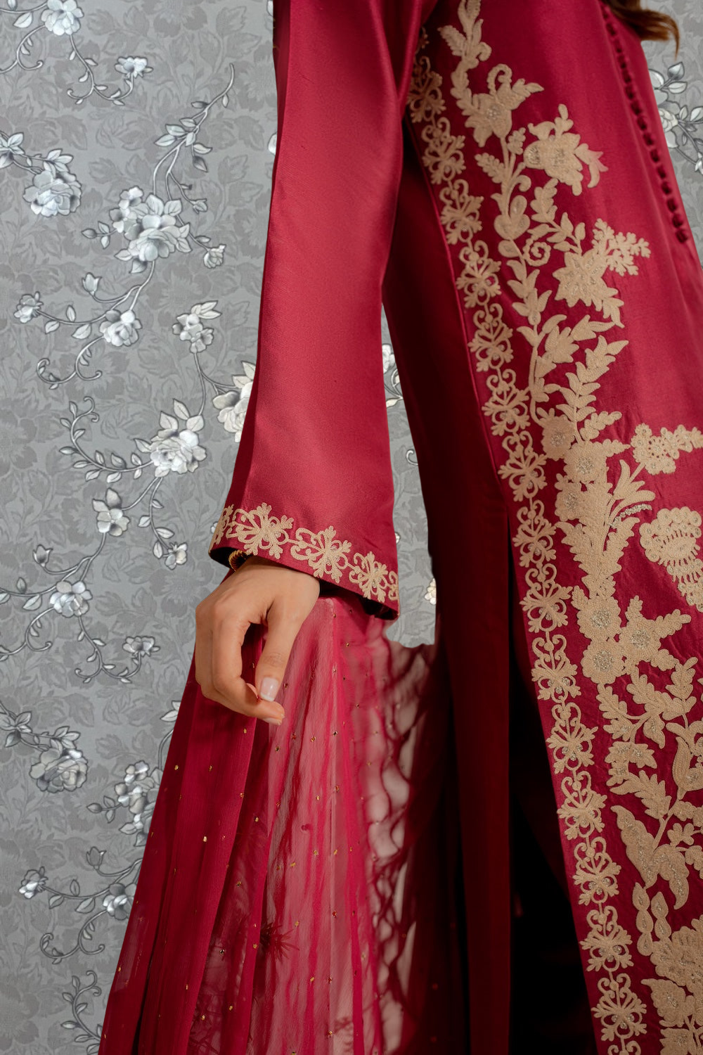 A close-up view of a person wearing a red and gold embroidered dress with a long sleeve, standing against a gray floral background.