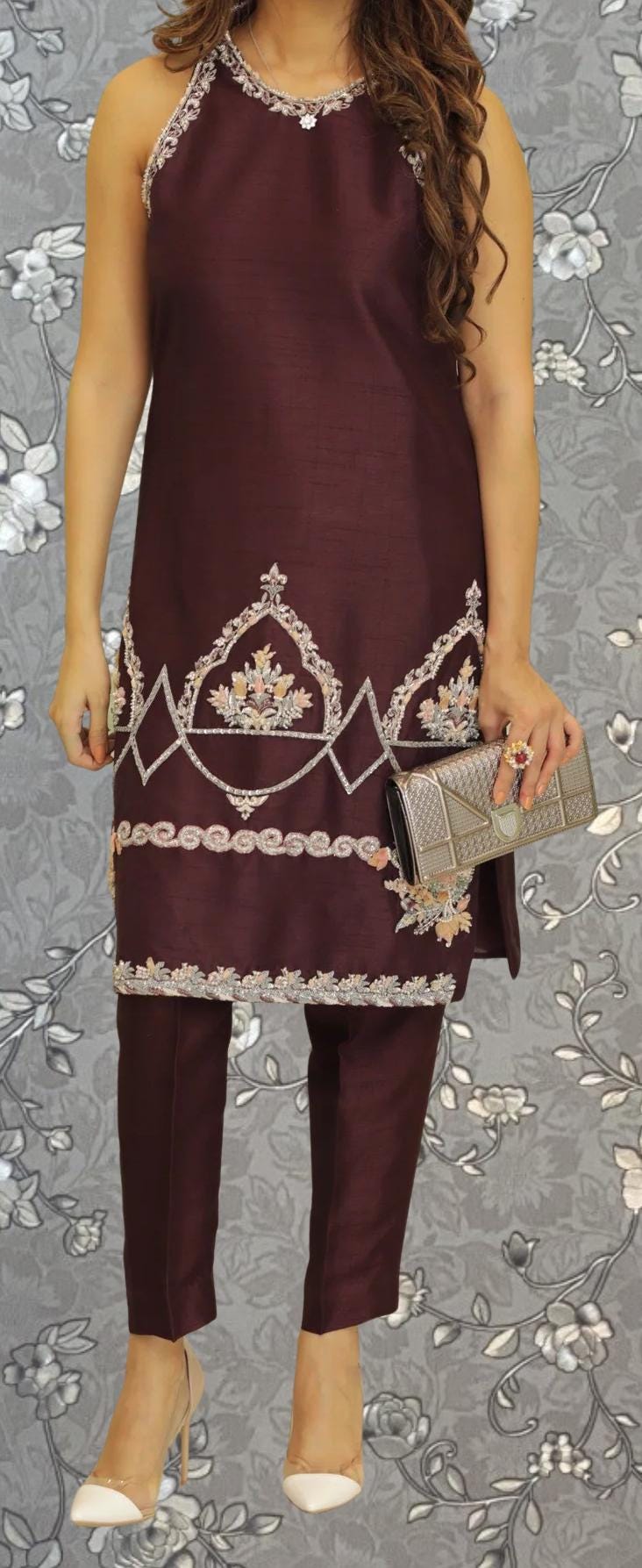 A woman wearing a burgundy dress with intricate embroidery and holding a small clutch purse stands against a gray floral background.