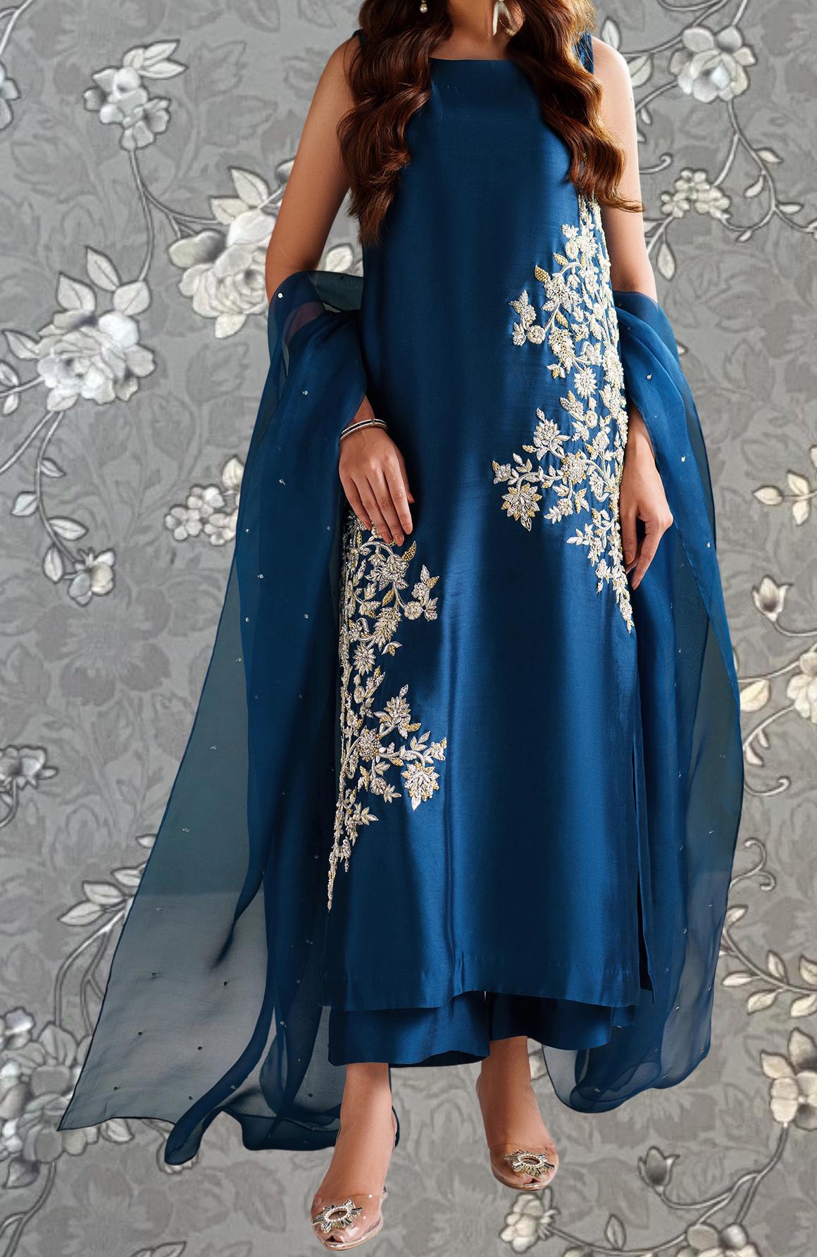 A woman wearing a blue dress with white floral embroidery, standing against a gray floral background.