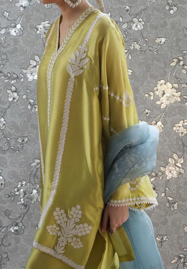 A woman wearing a yellow dress with white embroidered designs, standing against a gray floral background.