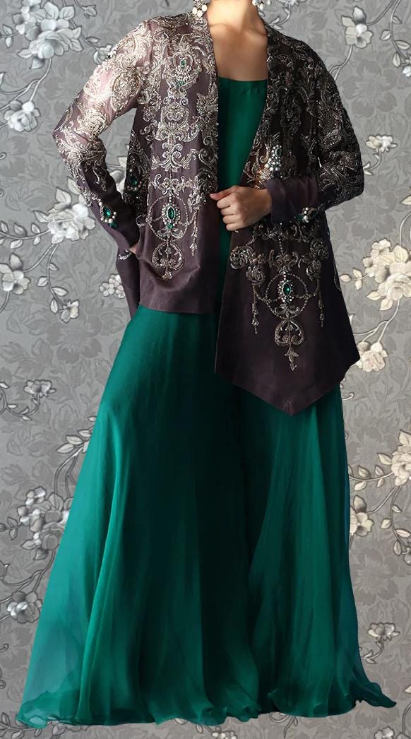 A woman wearing a long, green dress with a brown jacket over it, standing against a gray floral background.