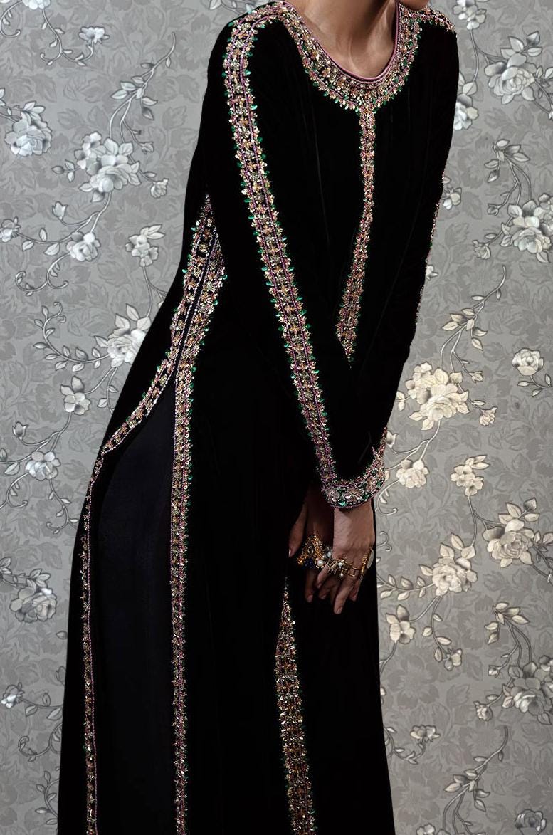 A person wearing a long black dress with intricate beaded and embroidered details, posing against a floral patterned background.