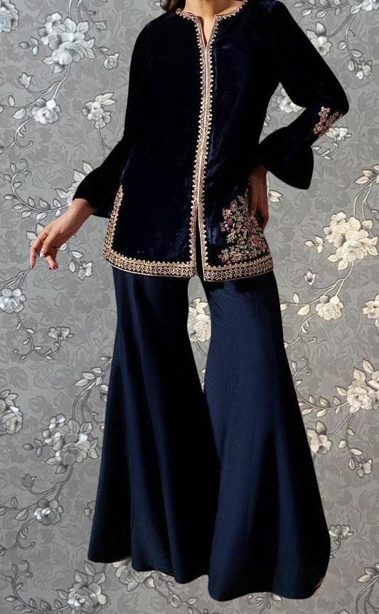 A woman wearing a black velvet dress with gold embroidery and a floral pattern on the skirt, posing against a gray floral background.