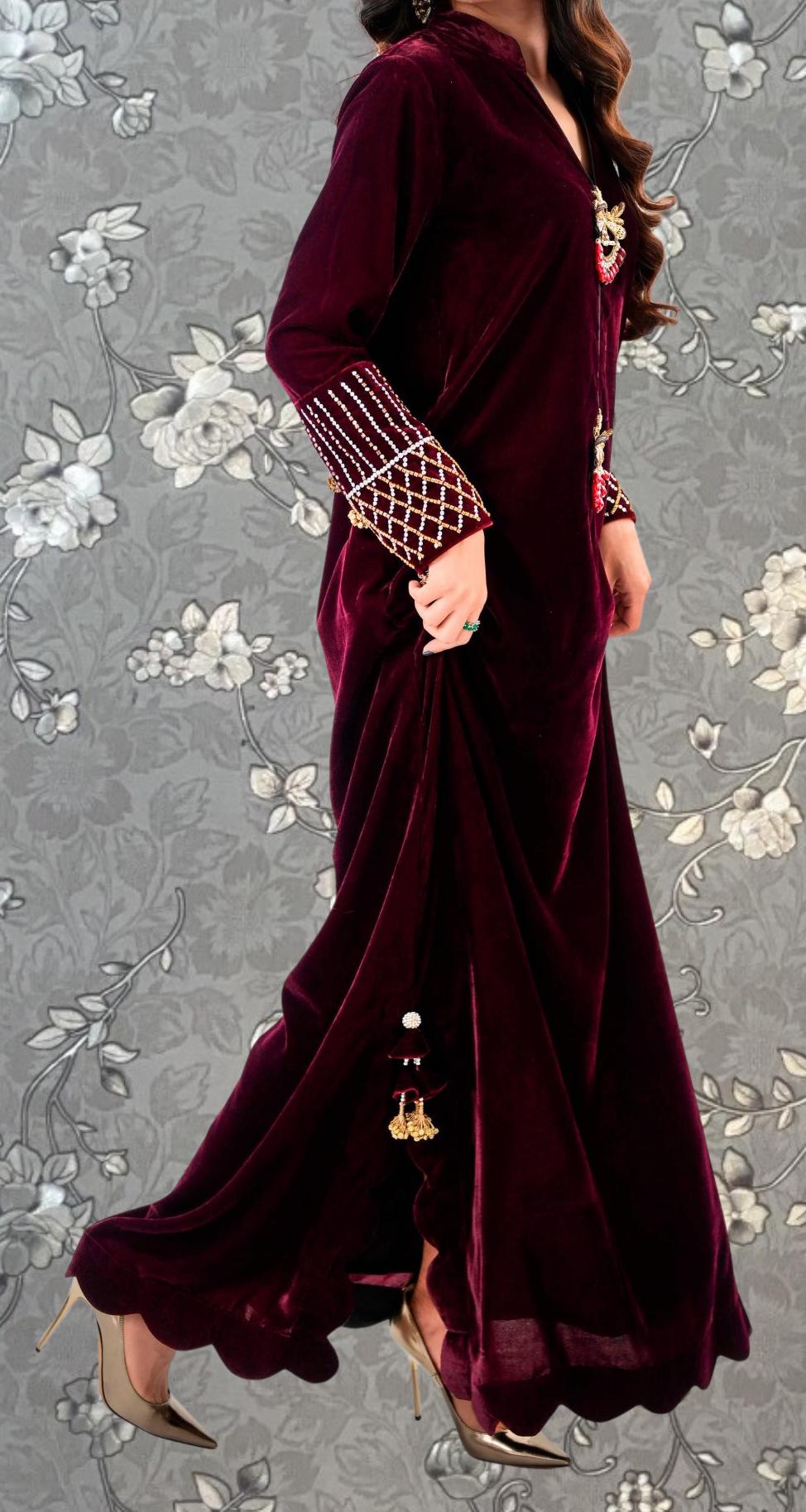 A woman wearing a long, burgundy velvet dress with intricate embroidery and gold accents, standing against a gray floral background.