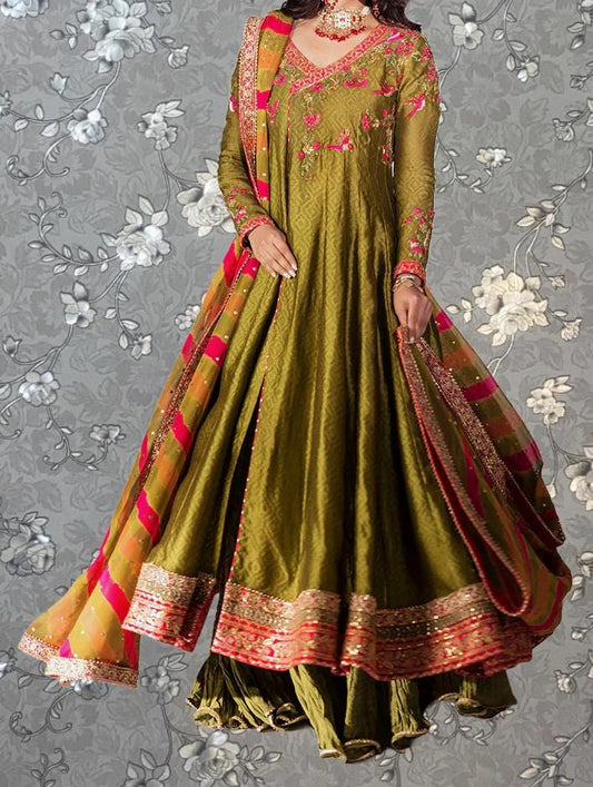 A woman wearing a long, green dress with intricate embroidery and a matching skirt, posing against a gray floral background.