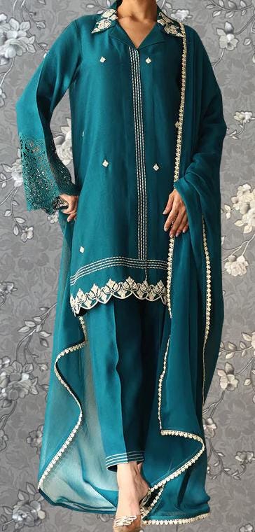 A person wearing a teal-colored dress with intricate embroidery and lace details, standing against a gray floral background.