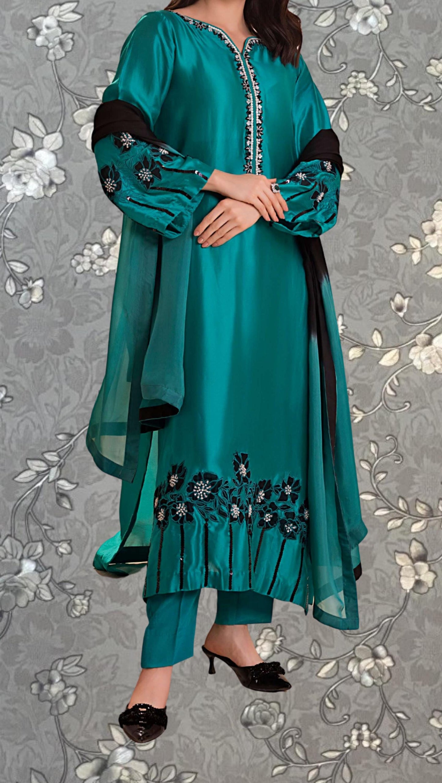 A woman wearing a teal green dress with floral embroidery and a black shawl, standing against a gray floral background.