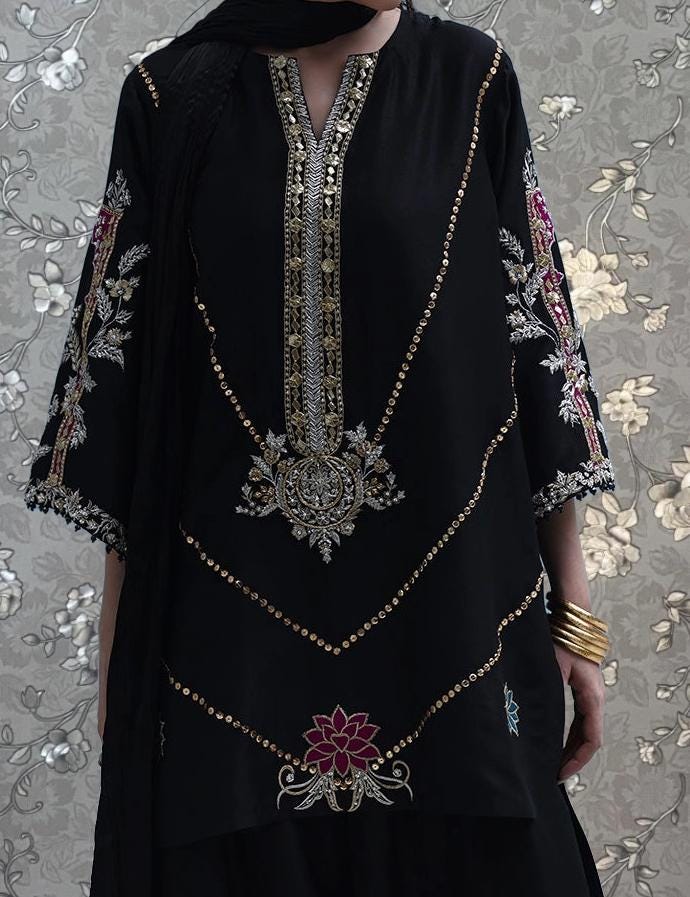 A person wearing a black dress with intricate gold embroidery and floral designs, standing against a floral patterned background.
