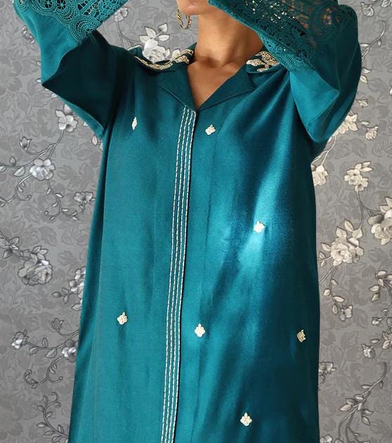 A person wearing a teal-colored dress with lace sleeves and small white flowers on the bodice stands against a gray floral background.