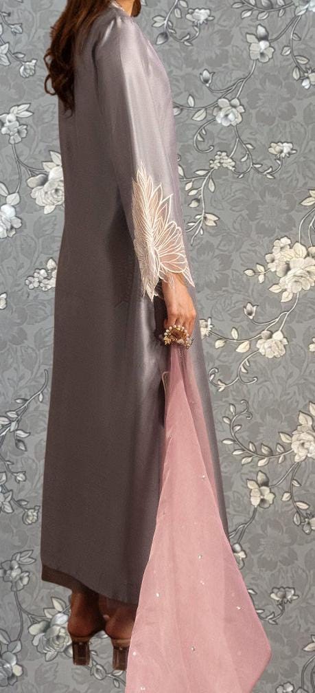 A woman wearing a long gray dress with a pink floral pattern on the sleeves, standing against a gray floral background.