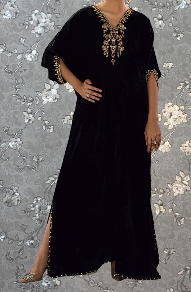 A person wearing a long black dress with gold embroidery stands against a gray floral background.