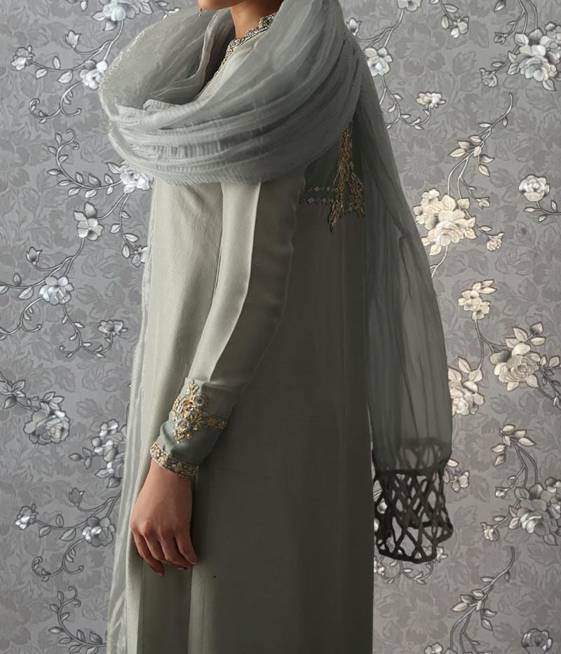 A person wearing a gray dress with a long, flowing scarf stands against a gray floral background.