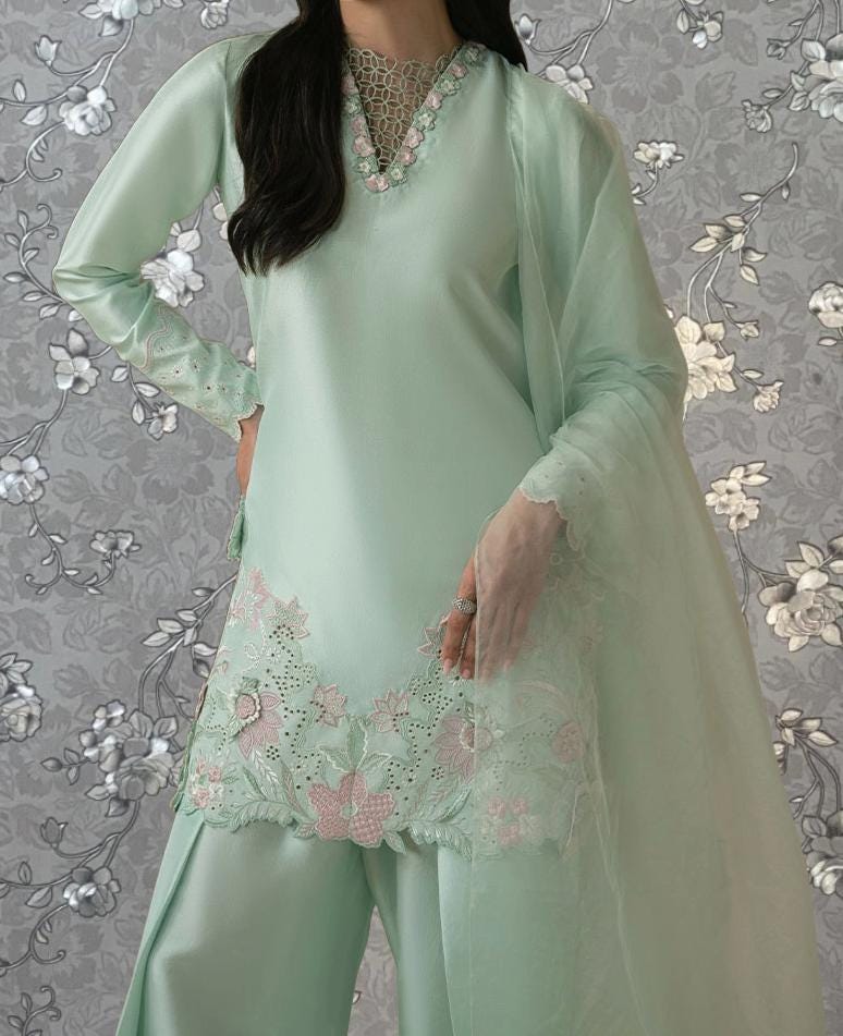 A woman wearing a mint green dress with intricate embroidery and lace details, standing against a gray floral background.