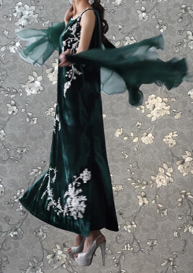 A woman wearing a long, green dress with white floral embroidery is shown in mid-jump, with her arms outstretched and her hair flowing behind her.