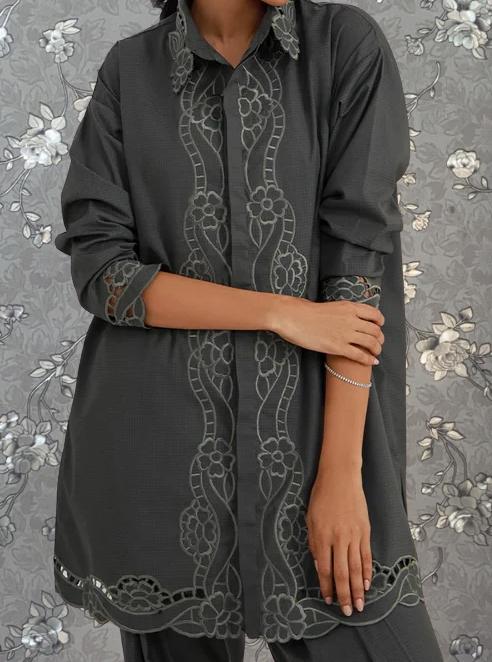 A person wearing a gray dress with intricate embroidered patterns on the sleeves and collar stands against a gray floral background.