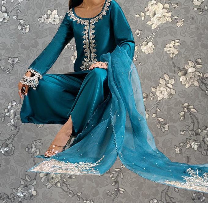 A woman wearing a teal blue dress with intricate embroidery and a matching blue shawl is sitting against a gray floral background.