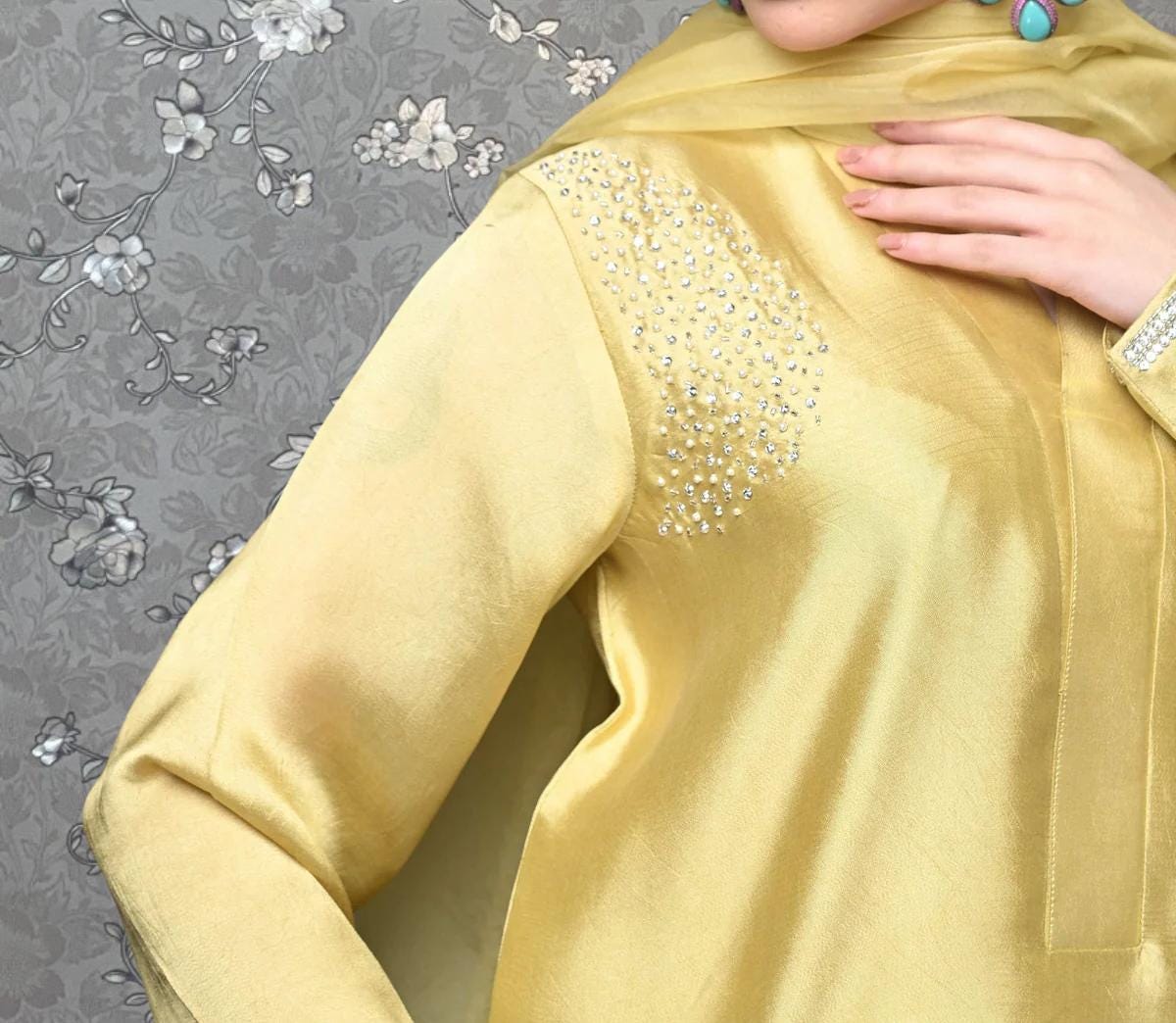 A close-up of a person wearing a gold dress with a floral pattern and a beaded embellishment on the shoulder.