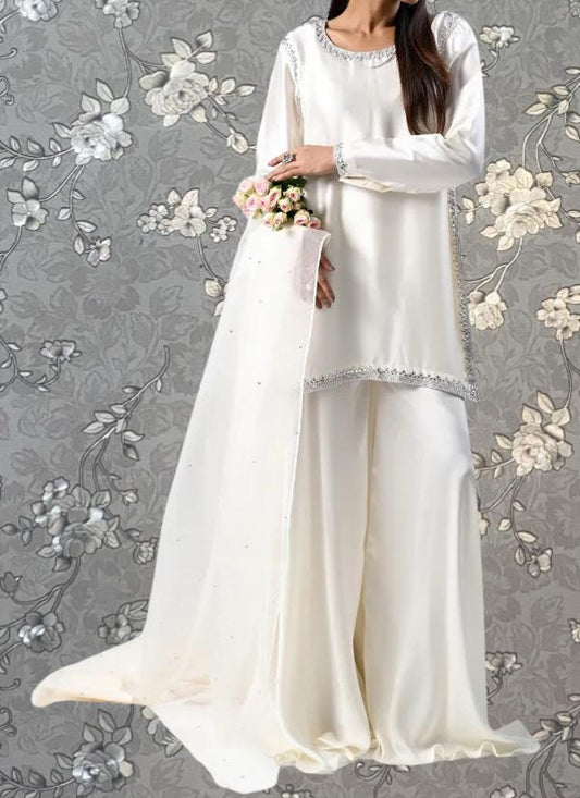 A woman wearing a long white dress with a long train, holding a bouquet of flowers, standing against a gray floral background.
