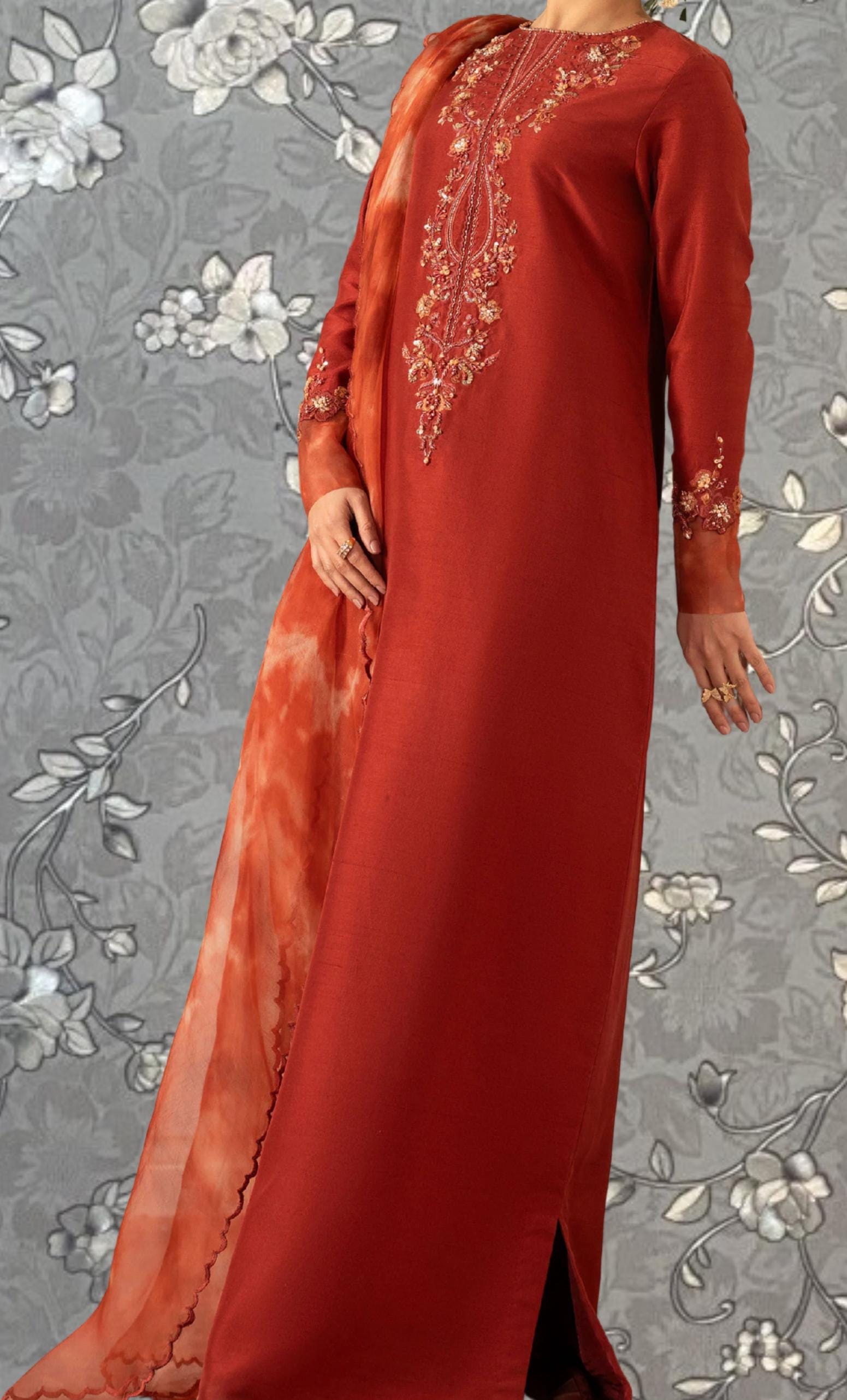 A woman wearing a long, red dress with intricate embroidery stands against a gray floral background.