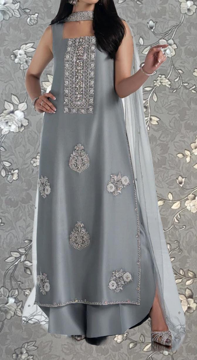 A woman wearing a gray dress with intricate embroidery and floral designs, standing against a gray floral background.